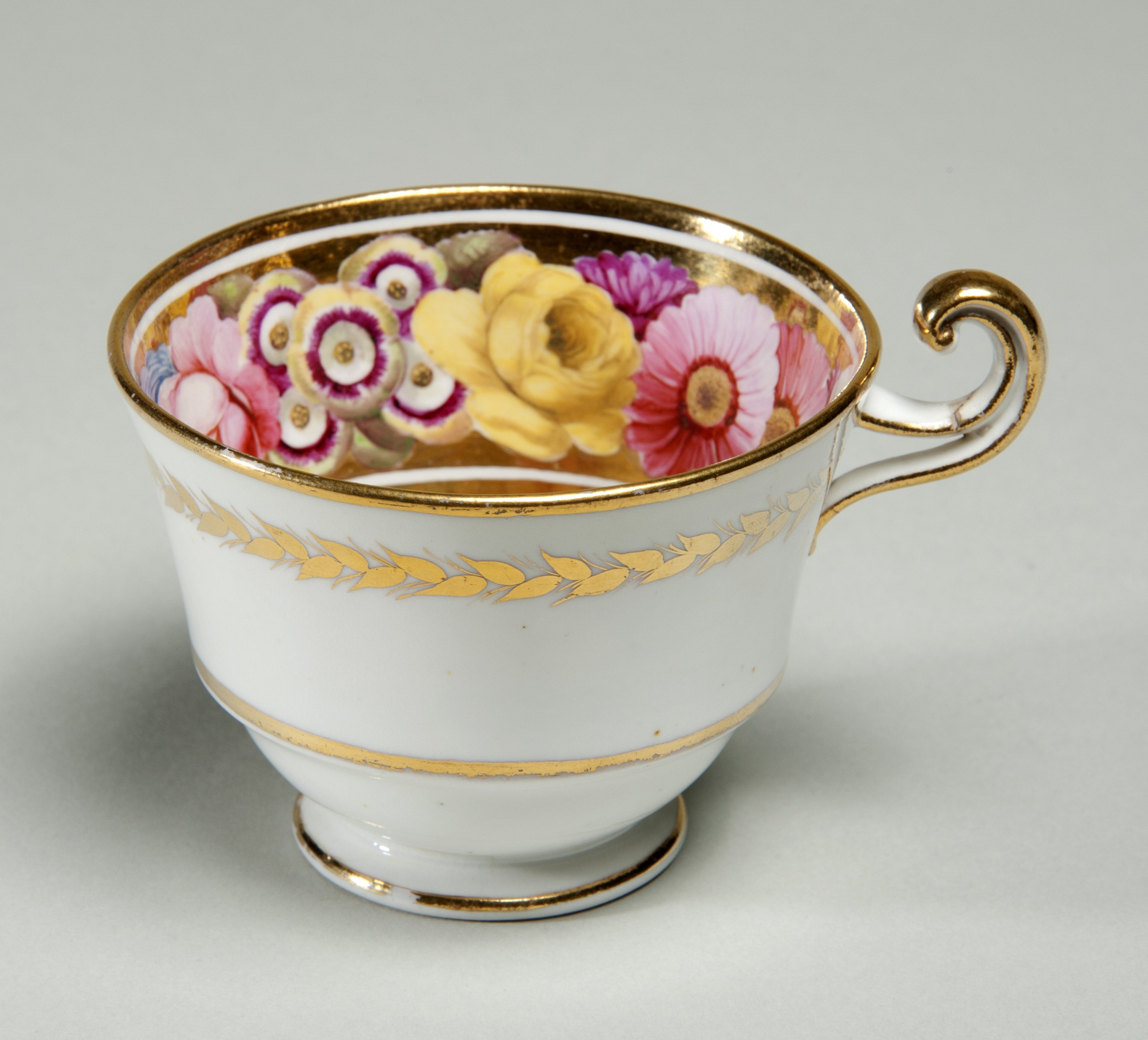 Colour photograph of a delicate looking teacup with pink and yellow flowers painted inside. It has gold edges