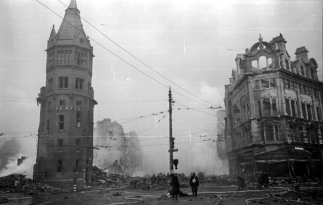 Black and White Photograph of a bombed street with a tower on the left and tall building on the right