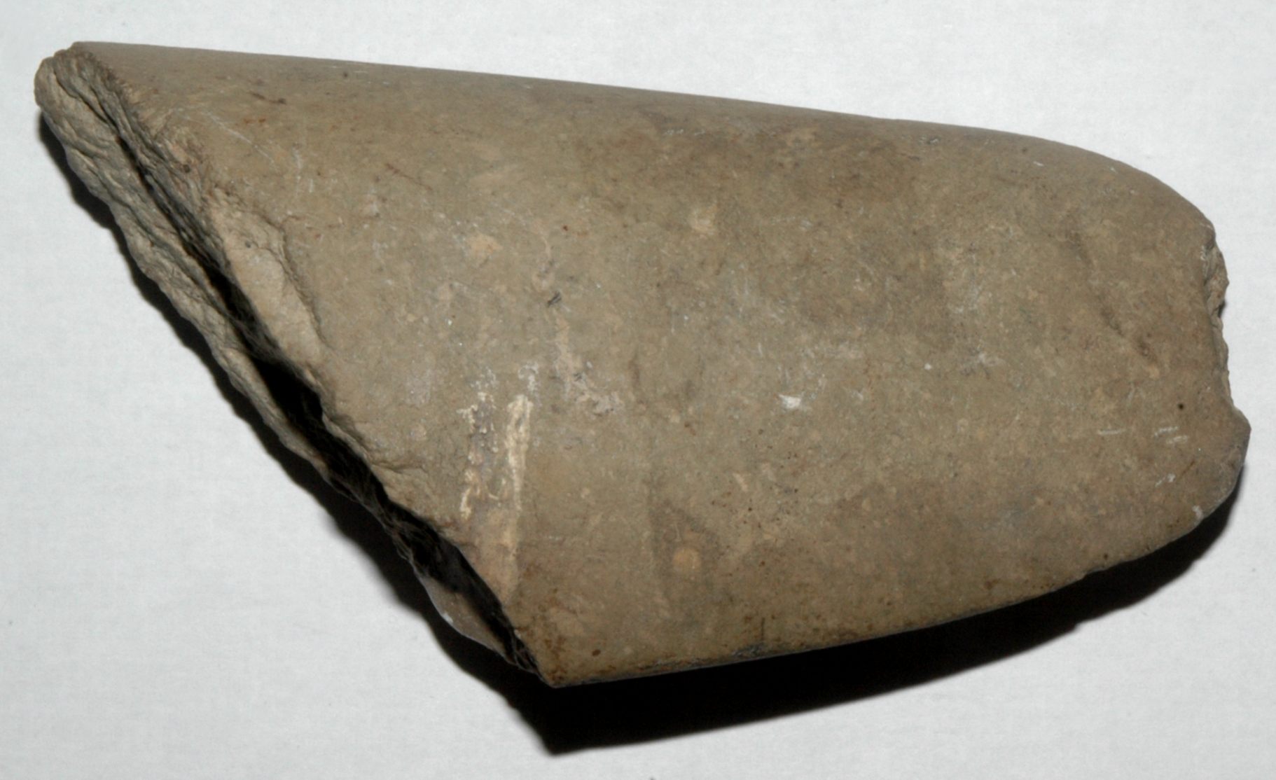A Neolithic polished stone axe, roughly triangular in shape with a smooth, slightly rounded cutting edge. The axe has a weathered, earthy surface, indicative of its age, and shows signs of careful craftsmanship with a tapered body. The axe is placed on a plain, light background, highlighting its texture and form.