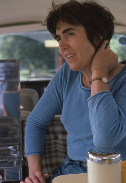 Maureen Roffey as a young woman with short dark hair, wearing a blue long-sleeved T-shirt, smiling while touching her neck. She is sitting on a grey checked seat in what appears to be a vintage vehicle, with a rear window behind her.