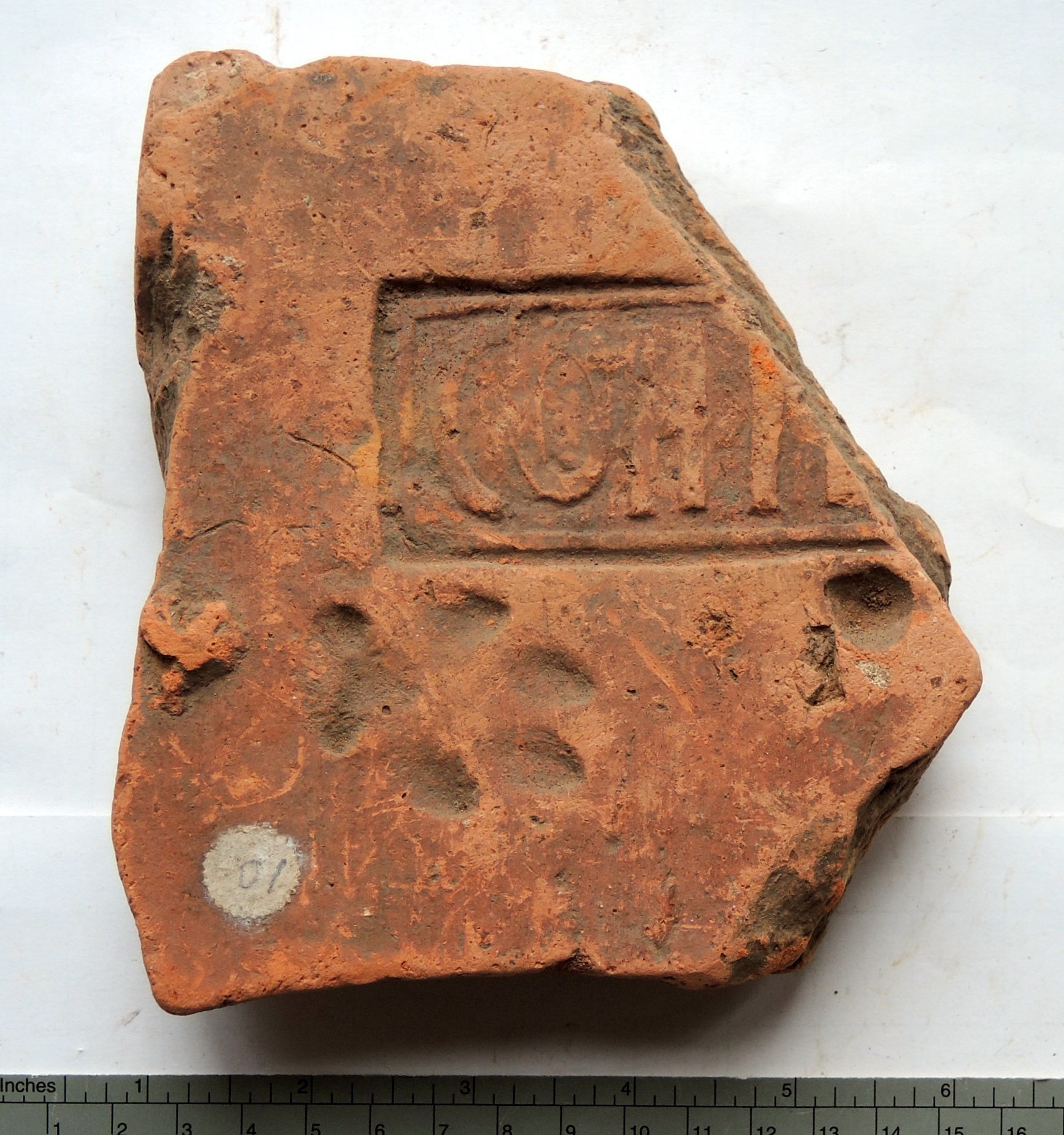A colour photograph of a broken Roman tile fragment made of reddish-brown terracotta. The surface features a rectangular stamped inscription in capital letters within a bordered frame. The tile is worn and irregularly shaped, with visible chips and cracks. An animal paw print is impressed below the inscription, likely made while the clay was still wet. A small circular museum label marked "07" is visible near the bottom left corner. A ruler along the bottom edge of the image provides scale in inches and centimetres.