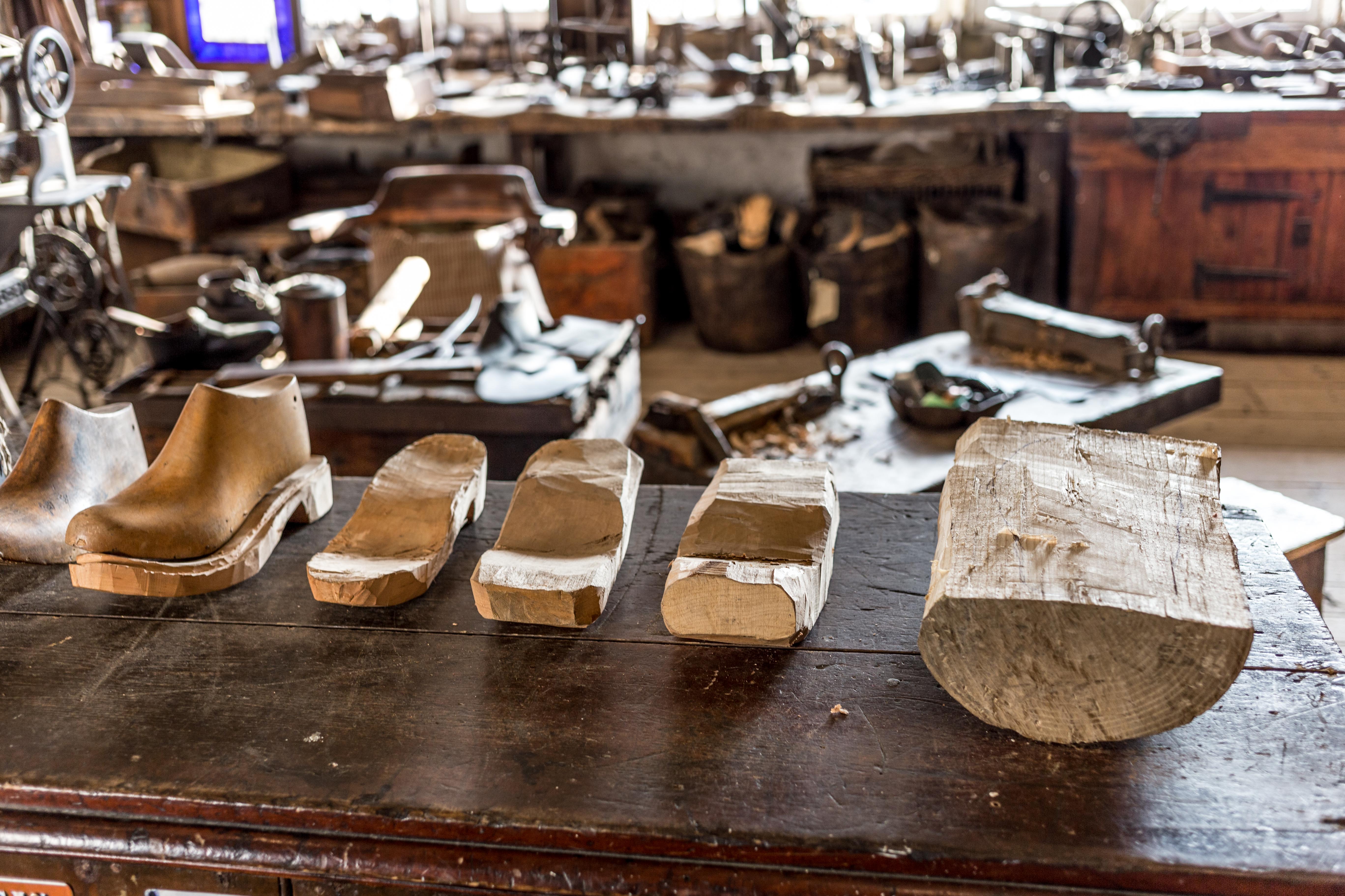 Colour photograph showing a raw lump of wood on the right hand side, and the finished clog on the left.  In between are three stages of the sole being carved.