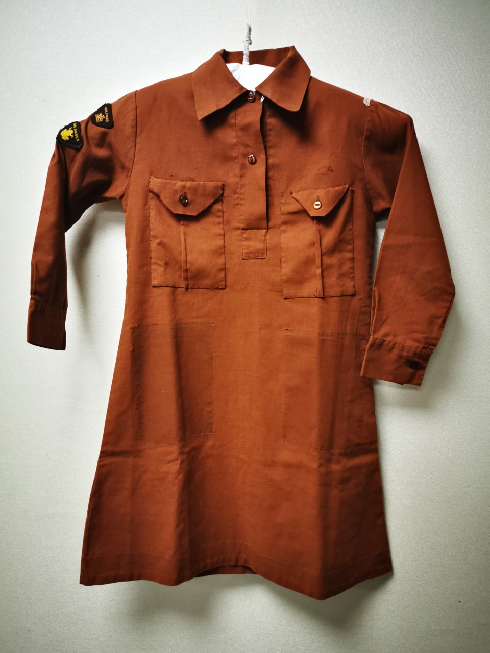 Colour photograph showing a brown shirt dress with collar and chest pockets. There are darker patches on the bottom half of the dress which show where the pockets used to be.