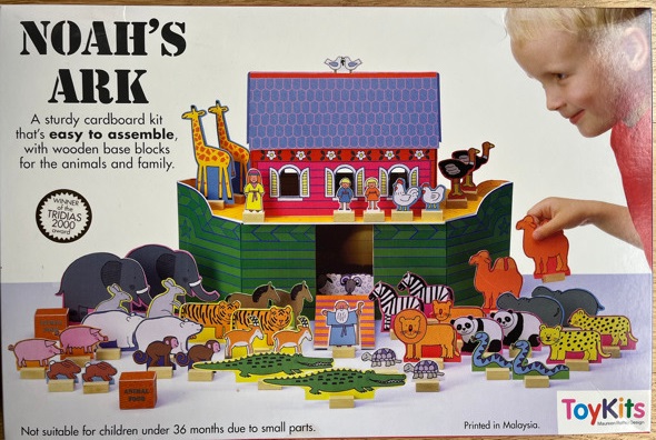 Packaging for a Noah's Ark kit created by Maureen Roffey. The packaging is white, with an image of a colourful cardboard Noah's Ark in the centre, with pairs of cardboard animals on top and alongside it. The animals include elephants, pigs, giraffes, rabbits, pandas, crocodiles, snakes, camels, ostriches, chickens, horses, lions, zebras, sheep, and tortoises. There is a cardboard Noah, little boy and little girl standing on the Ark, and the blue roof has a pair of white doves. A little boy with blonde hair is pictured playing with the ark, placing one of the camels down. He is the creator's actual grandson, Ben. The box reads 'Noah's Ark' in black capital letters, and underneath black text says 'A sturdy cardboard kit that's easy to assemble, with wooden base blocks for the animals and family.' Beneath this is a black circle, with text inside reading 'Winner of the Tridias 2000 award'. The bottom of the box reads 'Not suitable for children under 36 months due to small parts.' and 'Printed in Malaysia.'. There is a colourful logo for the company, Toykits, in the bottom right corner.
