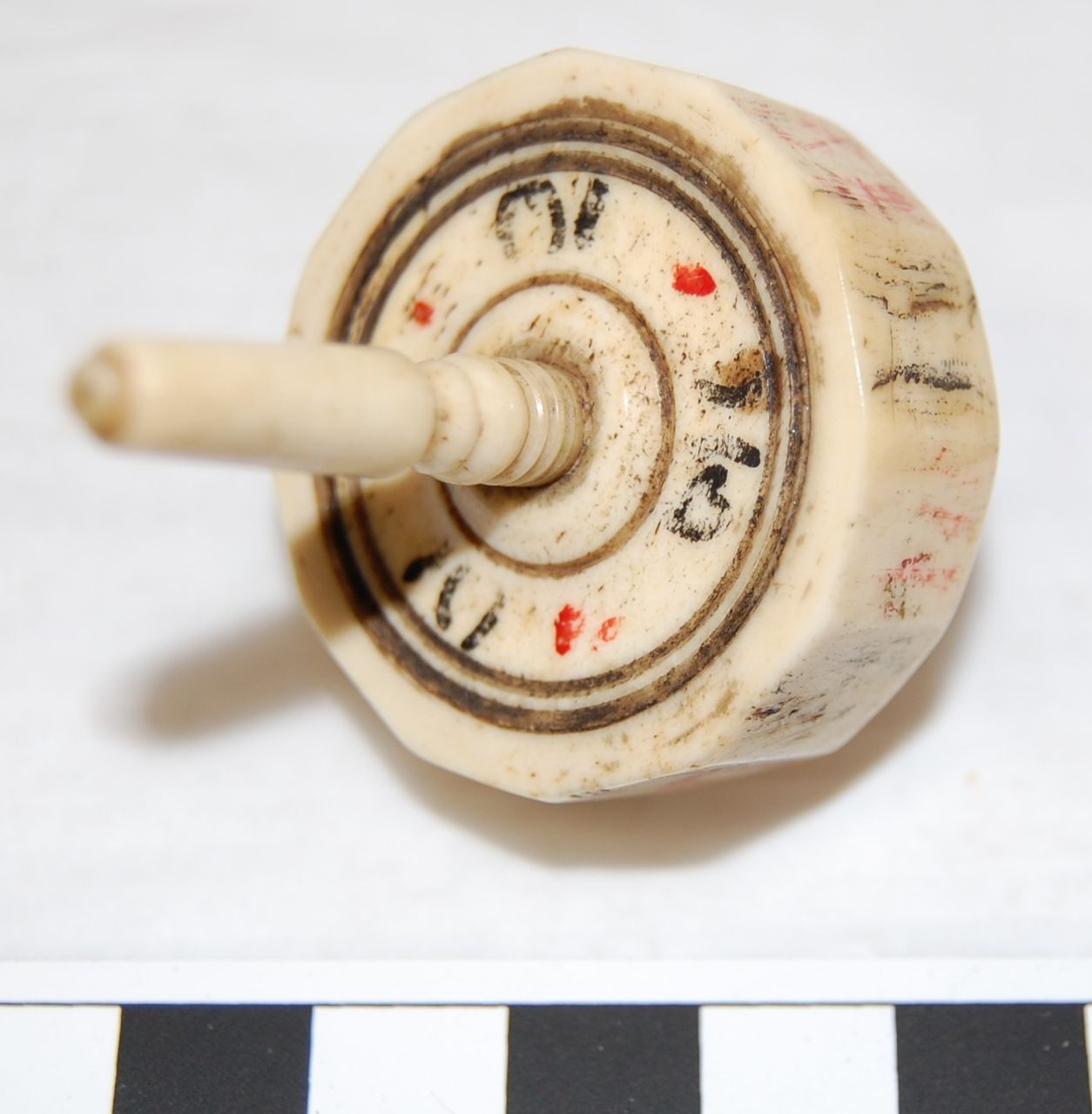 A vintage spinning top with worn markings, black and red symbols on a round, beige body. A ruler indicates scale.