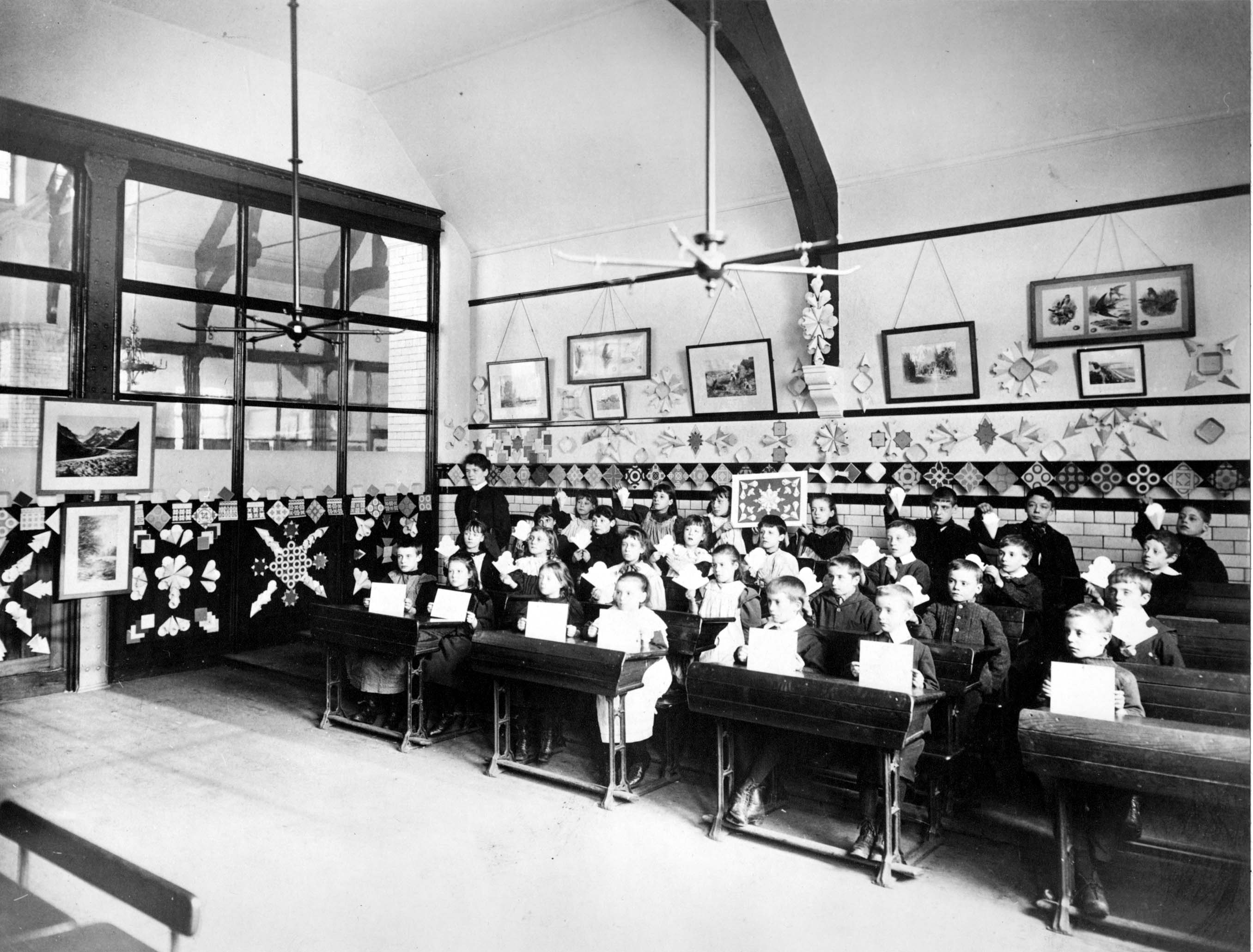 Black and white image of a school classroom around 1900. The pupils sit at paired desks in rows. The boys wear dark coloured button up jackets and the girls wear white aprons. Each child is holding up a piece of card with their artworks on. The room has a vaulted ceiling, and glass windows to the internal corridor beyond. The walls of the classroom are tiled and decorated with children’s artwork.