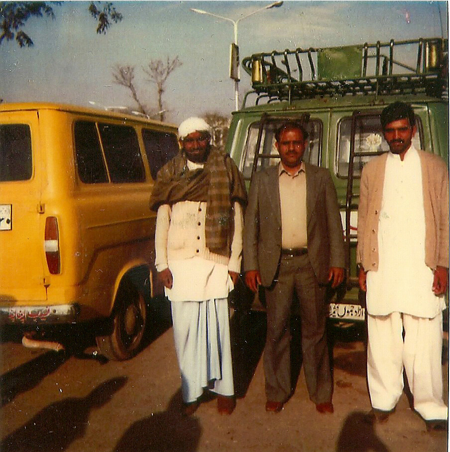 Colour photograph showing three men standing at the back of a green van.  The man in the middle is wearing a western style suit,  The men either side are wearing traditional Pakistani dress.