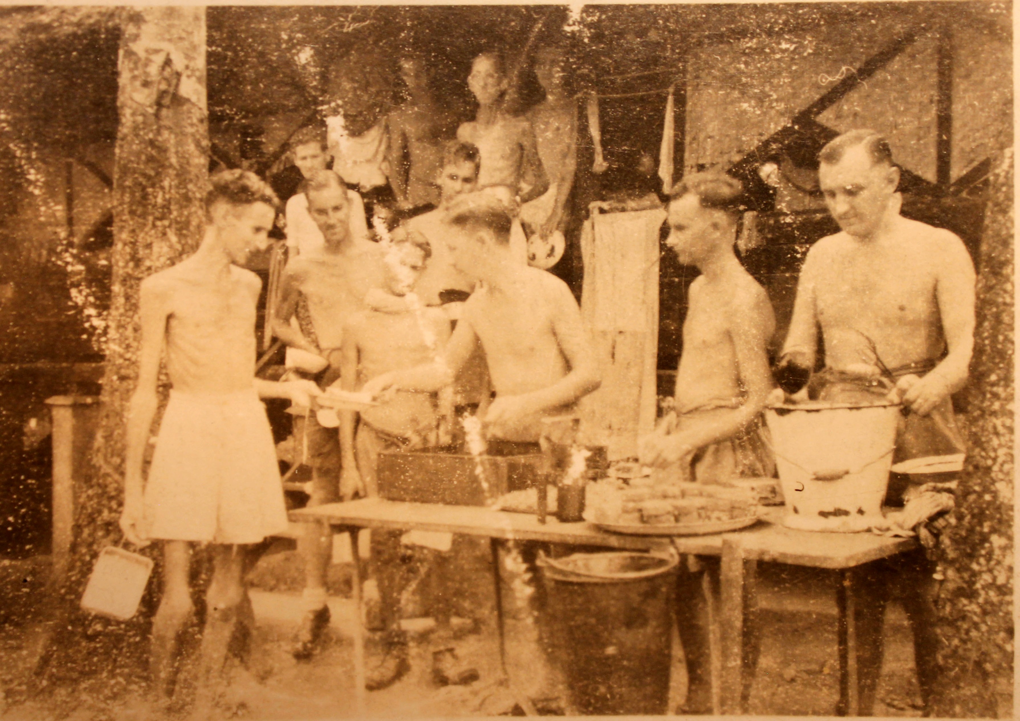 Photograph showing a line of men waiting to be served food. They are all only wearing shorts. Some are very underweight.