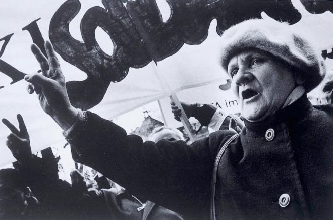 An elderly woman in a hat is holding up two fingers in the victory sign, with a large banner behind her. She is on a march.