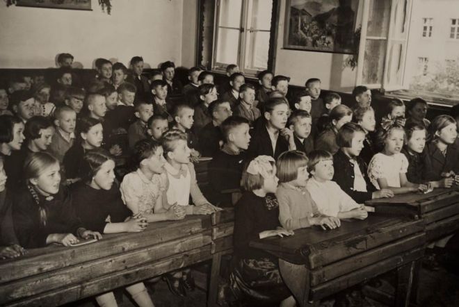 A crowded classroom with school children of varying ages sitting in lines at wooden desks.
