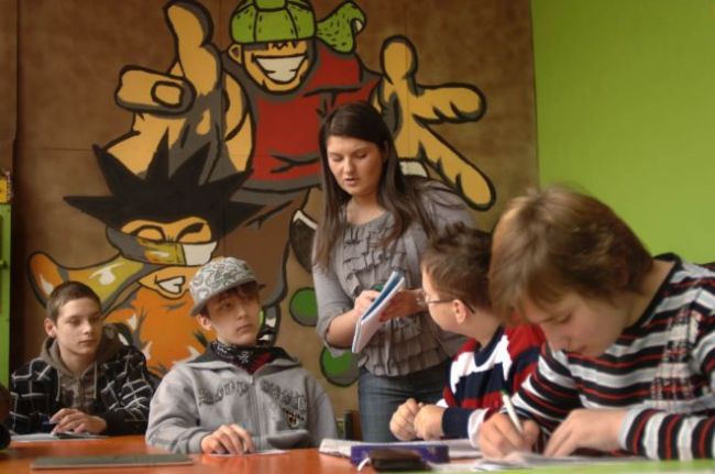 Four teenagers in casual dress sitting at desks listening to a teacher.