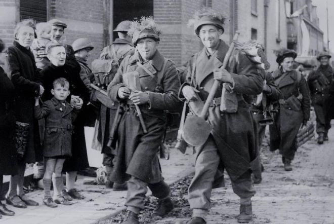 The Allied troops are smiling and there are men women and children on the pavement watching them go by.  The bystanders look quite happy.