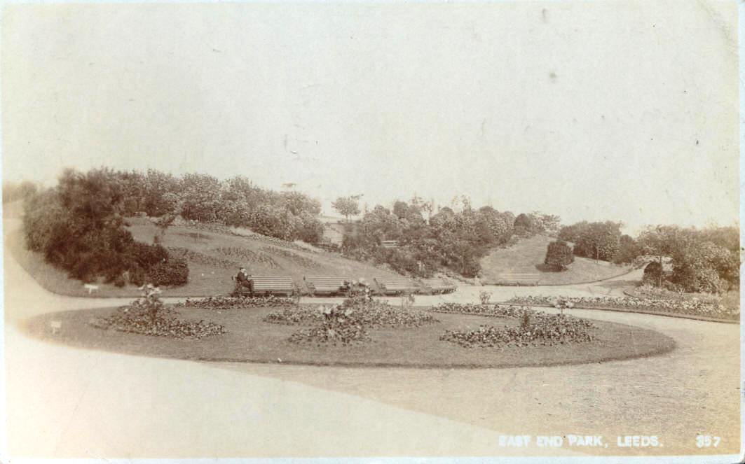 Faded black and white photograph of formal gardens. The centre of the image is a large, circular grassed island, with four ornamental kidney flower beds around the outside and a one circular one in the middle. This is surrounded by paths, with hillocks and shrubs in the background. There are benches around the outer edge and a man sits on one.