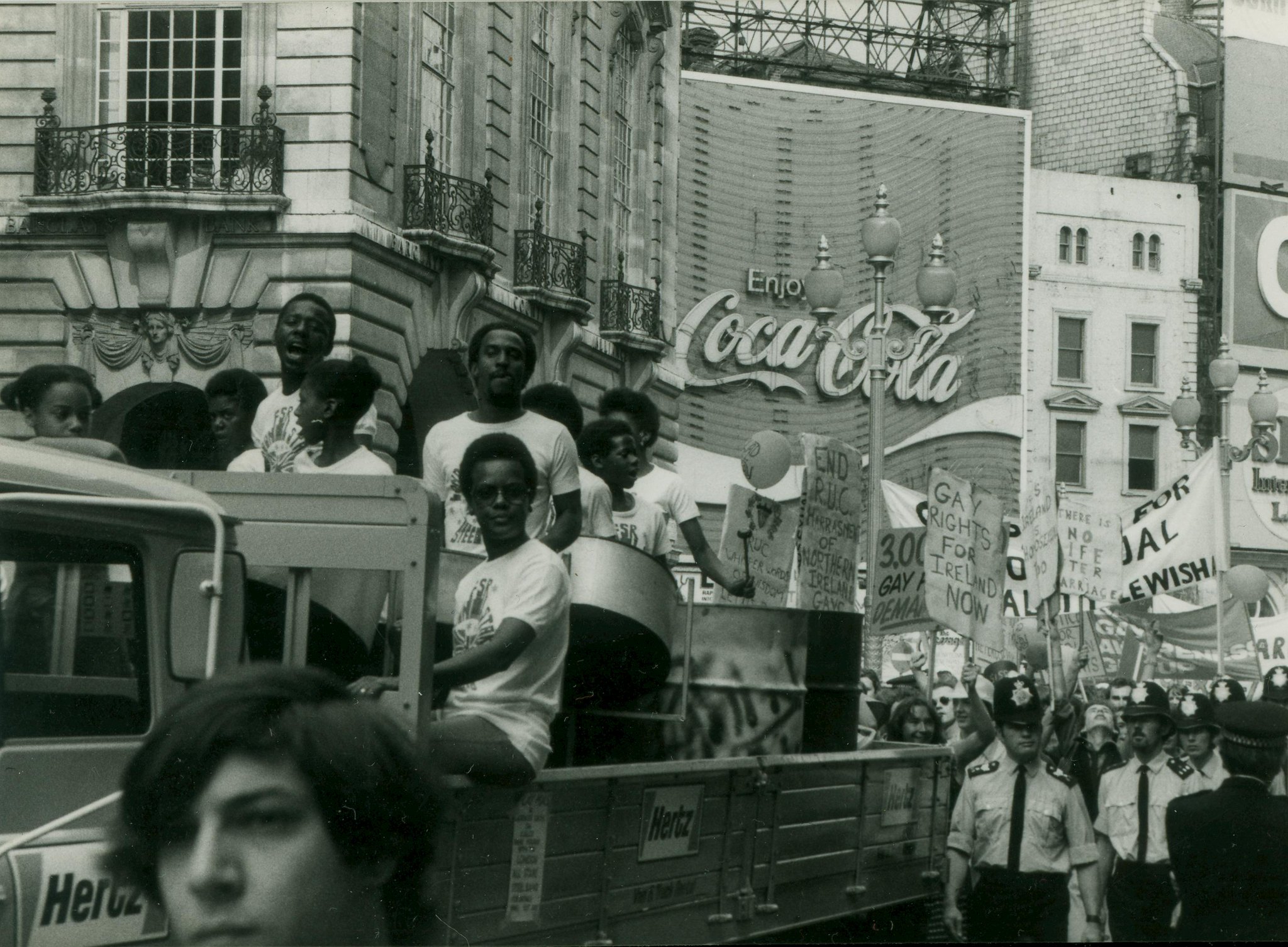 Black and white photograph of people on a float, playing drums. Police officers are walking beside the float.