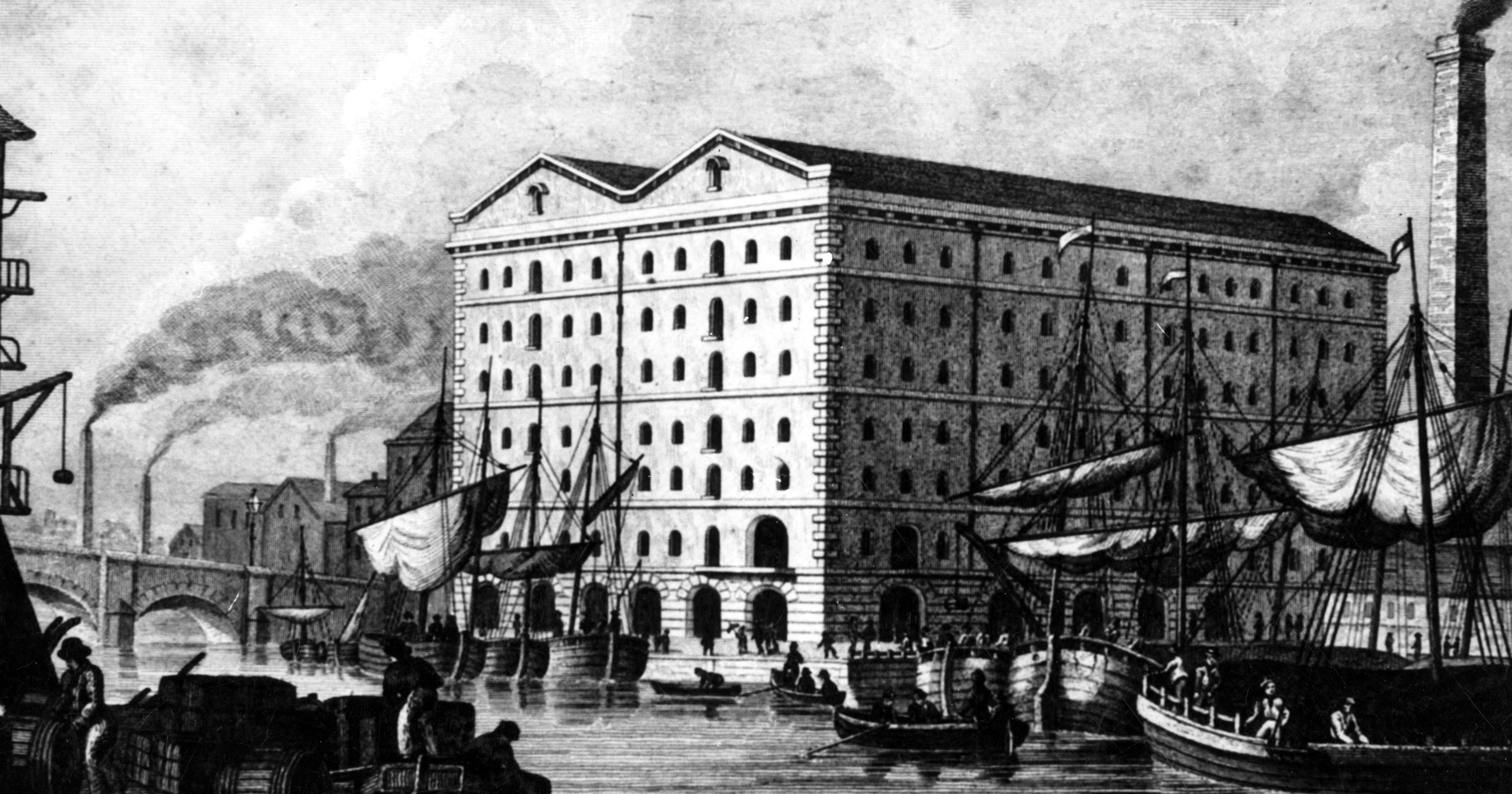 A black and white image of a large building with hundreds of window. Sailboats in the foreground. Large chimneys with smoke billow out in the background