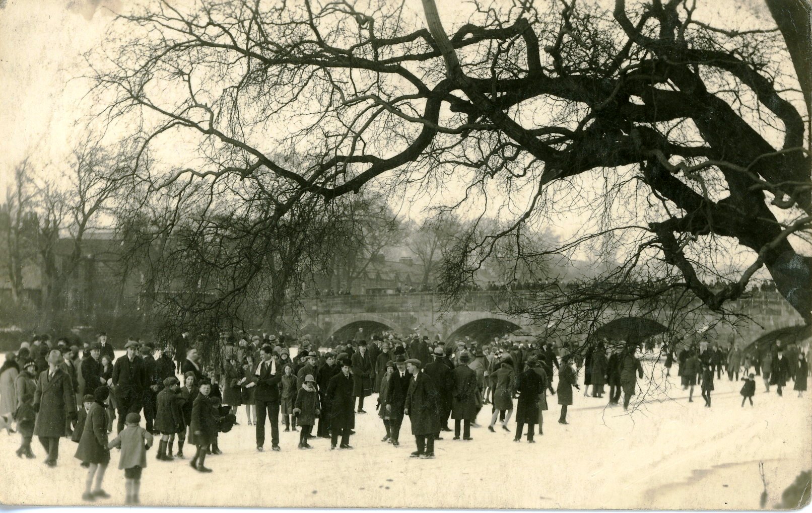Black and white photograph of many people (men, women and children) standing and playing on the frozen river Wharfe a Otley. The stone Otley bridge is in the background, alongside houses on the far bank