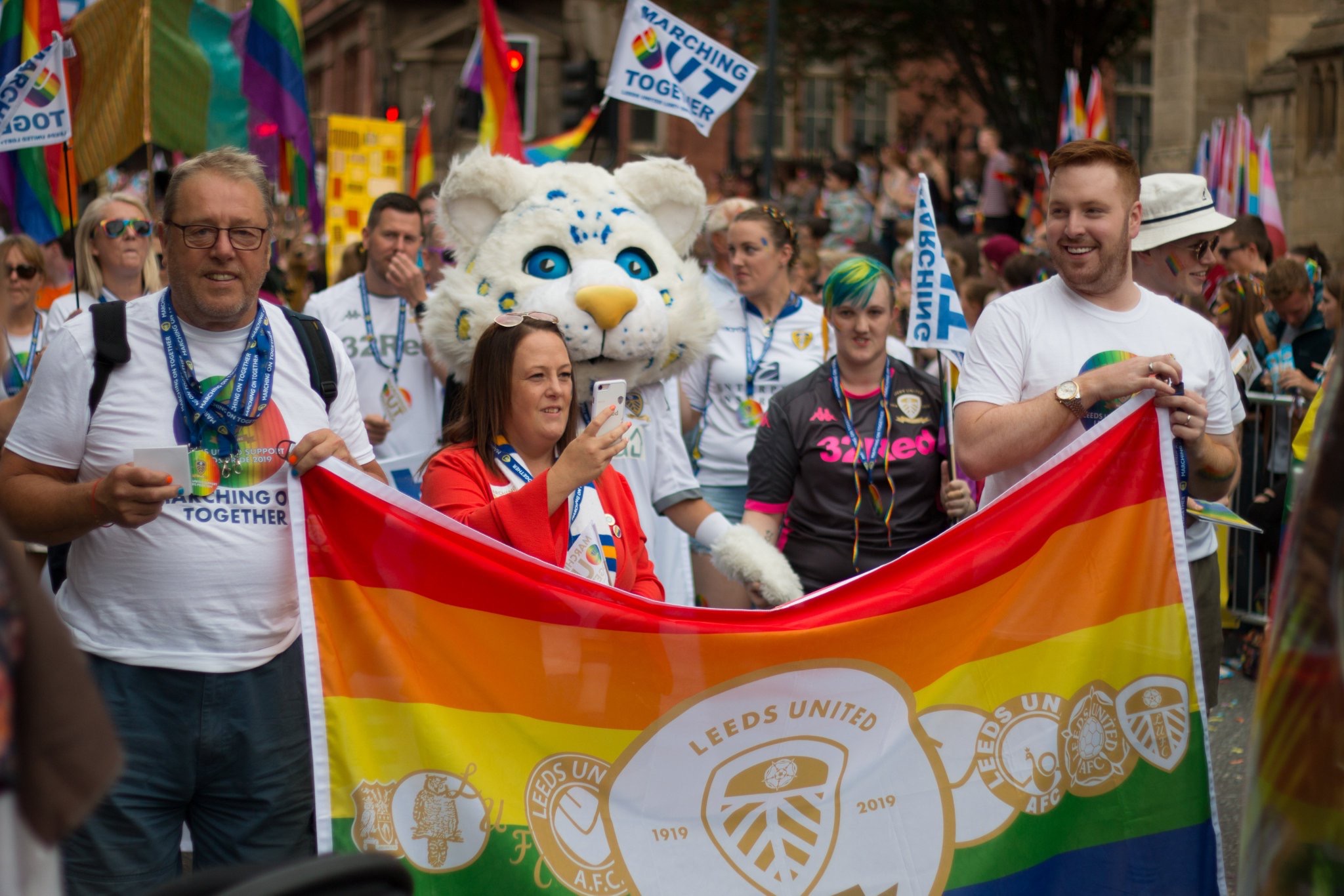 Colour photograph showing a crowd of people marching in the city of Leeds.  At the front several people are holding a large rainbow Leeds United flag