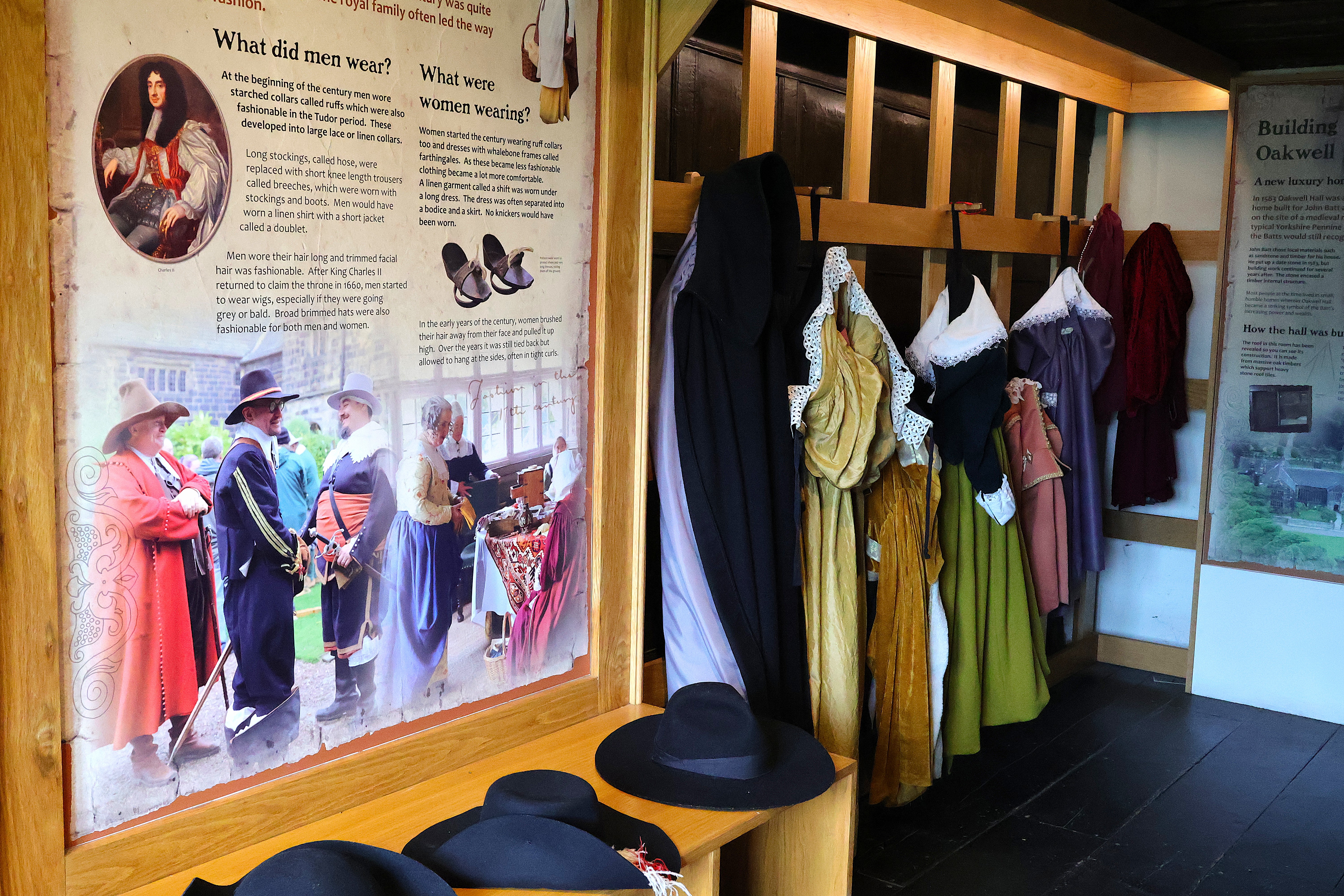 A historical museum exhibit showcases vintage clothing with hats, dresses, and cloaks on display, ready to be tried on by visitors. Informational panels describe period attire for both men and women.