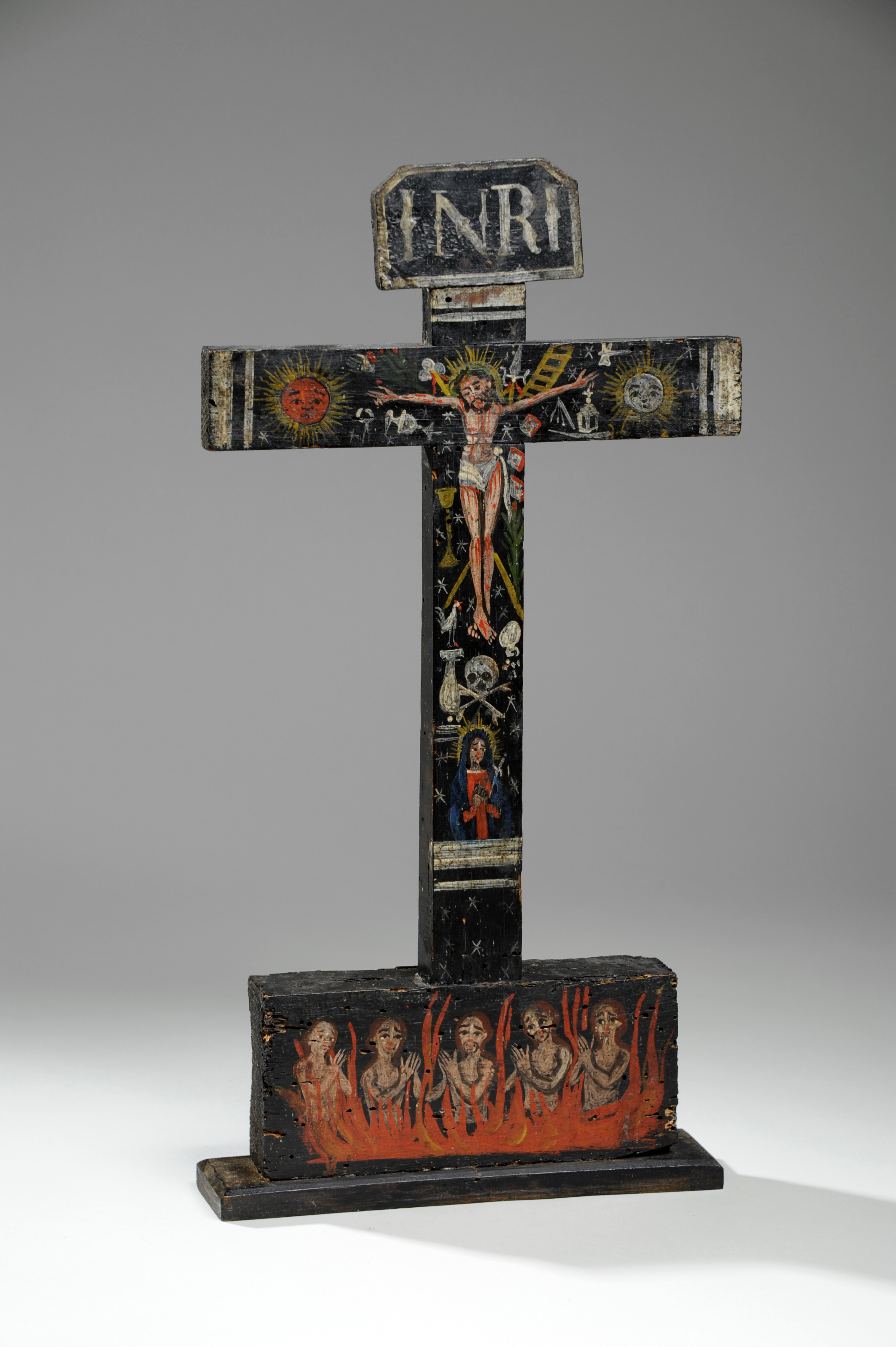 A painted wooden crucifix in a folk art style, featuring the crucified Jesus at the center with a golden halo and the inscription "INRI" above his head. Surrounding Jesus are symbolic elements including the sun, moon, stars, a ladder, and skull imagery. Below the crossbar, a depiction of the Virgin Mary or a saint is painted, while the base shows six anguished, nude figures with raised hands engulfed in stylised red and orange flames, symbolising purgatory or hell. The entire piece is painted on a black background with white star motifs and bordered in distressed wood tones.