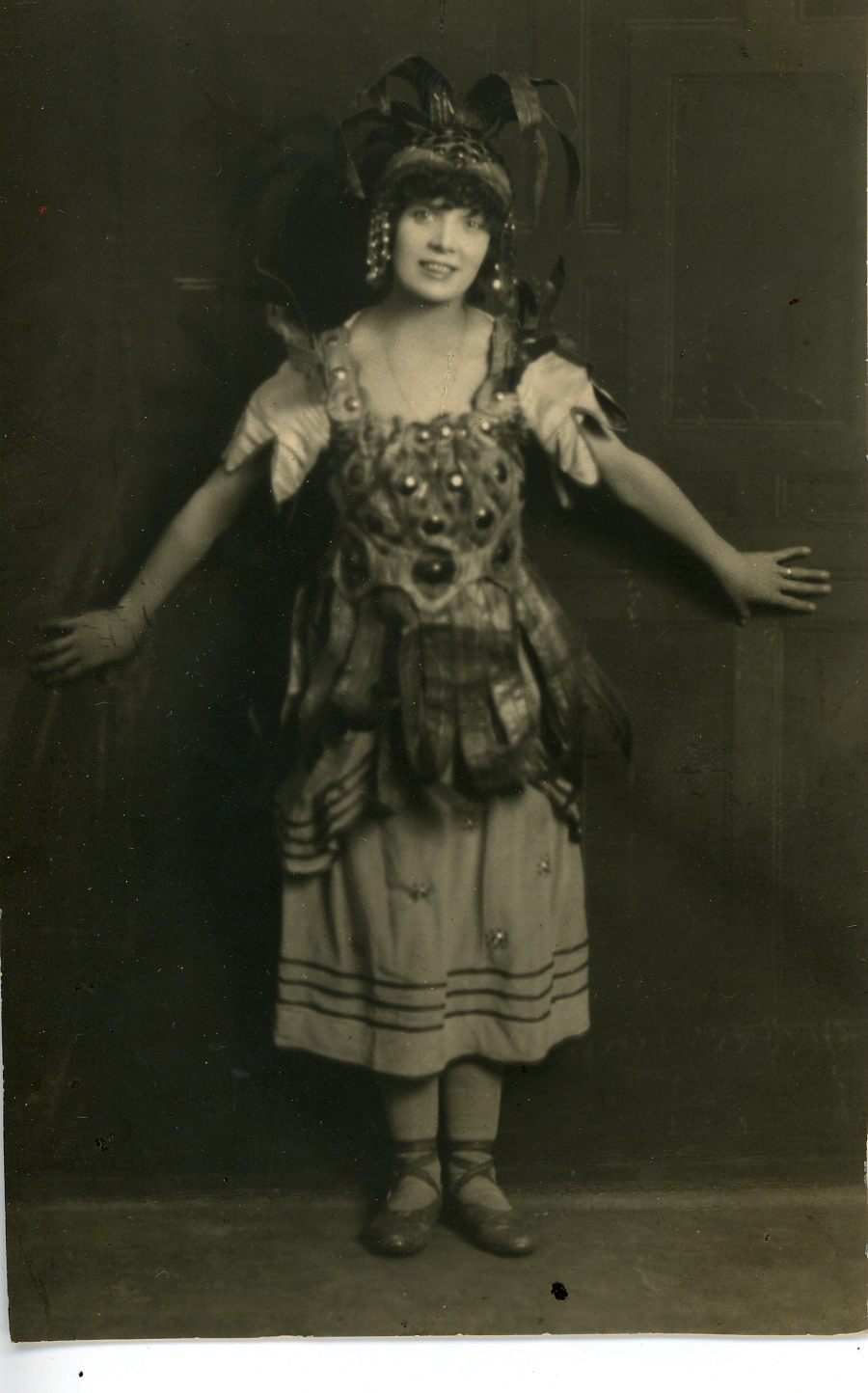 Black and white photograph showing a female actress in a pantomime costume. She is wearing a big headress and a dress with big, shiny button like decorations.