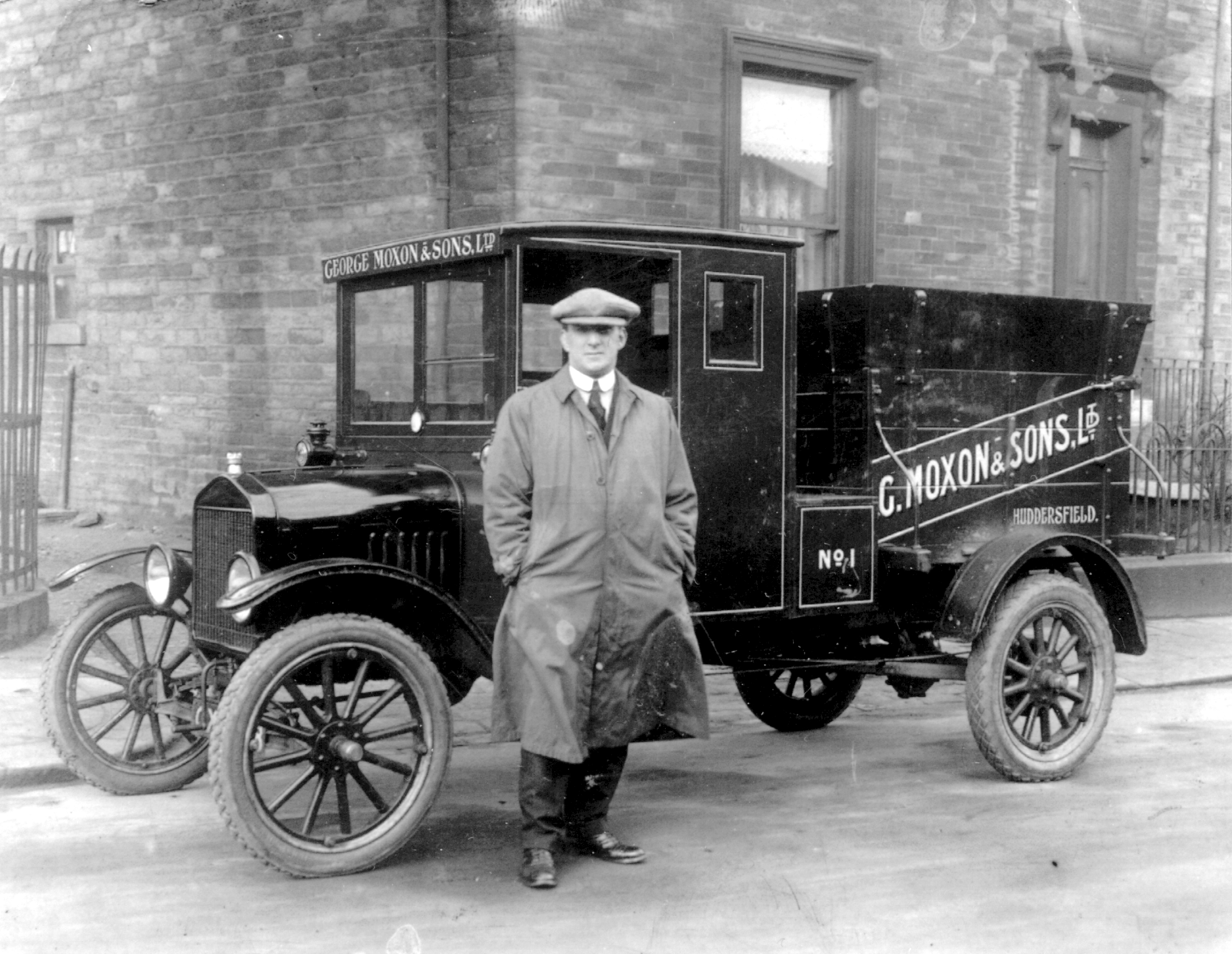 A black and white photograph depicting man in a trench coat and cap standing proudly beside an early 20th-century delivery vehicle with "C. Moxon & Sons Ltd" written on the side.