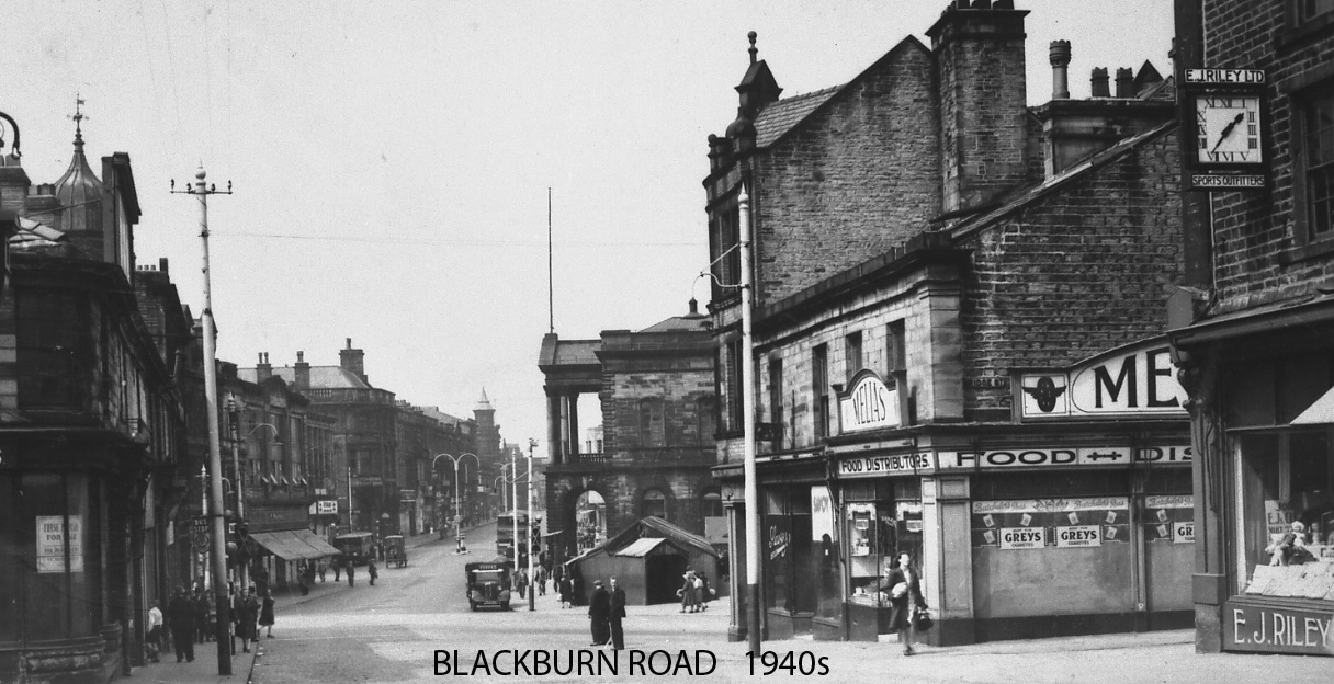Black and white photograph showing a wide road with many shops.  There is a bus in the distance and a car on the street.