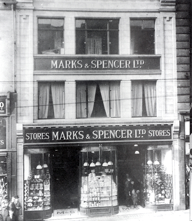 A formal black and white group portrait from around 1900 showing a lineup of young female staff at a Marks & Spencer Penny Bazaar in Cross Arcade. The women wear dark dresses with high collars and are positioned behind a wooden balustrade. Two men—likely managers—stand at the back, one wearing a uniform cap. The group is posed in front of an arched window in a stone building.