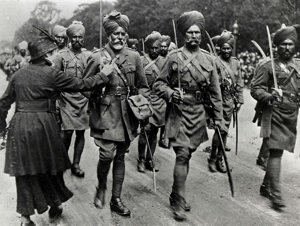 Black and white photograph showing Sikh troops wearing turbans and holding long swords. A woman is holding out flowers to one of the soldiers.
