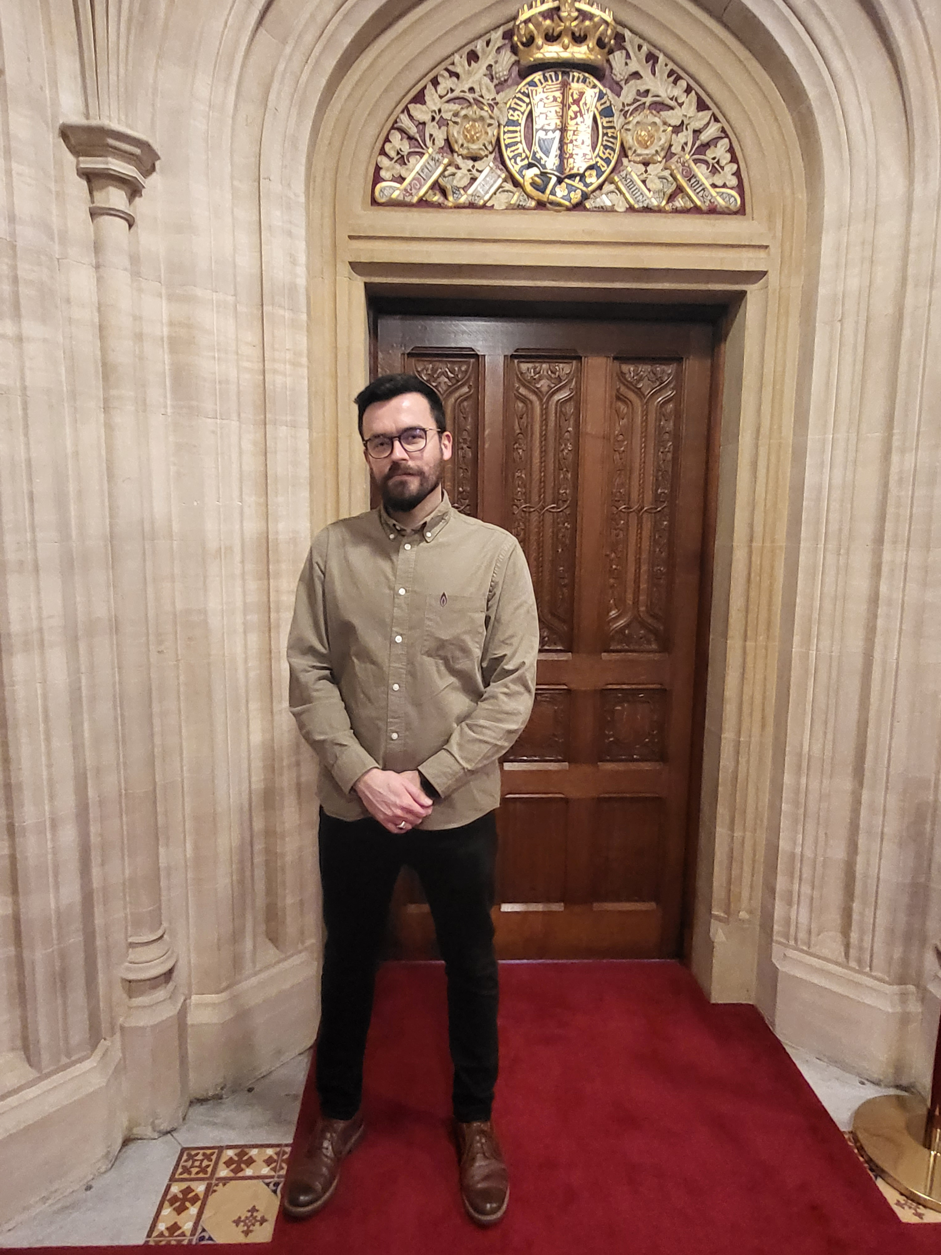 A  colour photograph of a man standing indoors in front of an ornate wooden door. The man has short, dark hair, a beard, and is wearing glasses. He is dressed in a light brown button-up shirt and dark pants, with brown leather shoes. The setting appears to be a grand, historical building, as the door is framed by intricate stone archways and adorned with a colorful coat of arms above it. The man stands on a red carpet, with his hands clasped in front of him, looking calmly at the camera.