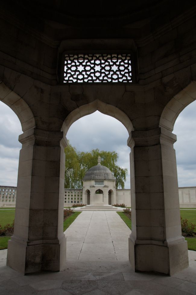 Photograph is looking through two pillars towards a similar structure.  There is grass on either side.  Looks like a circular enclosed area with a wall around