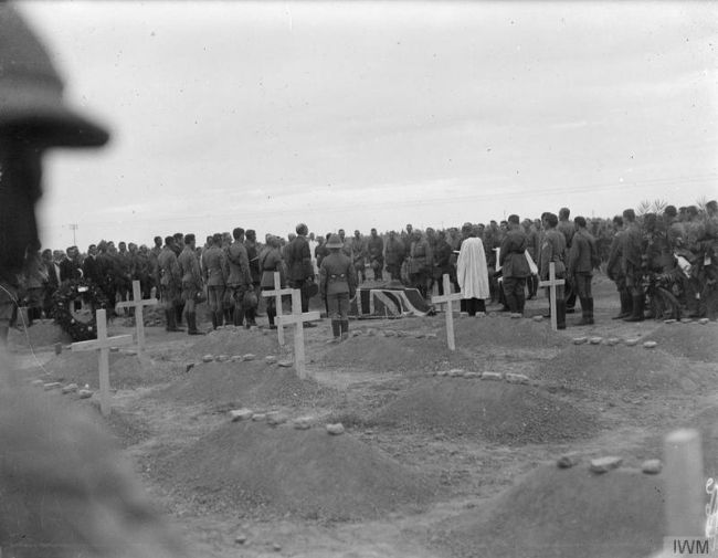 Black and white photograph of a rows of freshly dug graves with wooden crosses and many military personnel standing round a coffin draped in what looks like the Union Jack