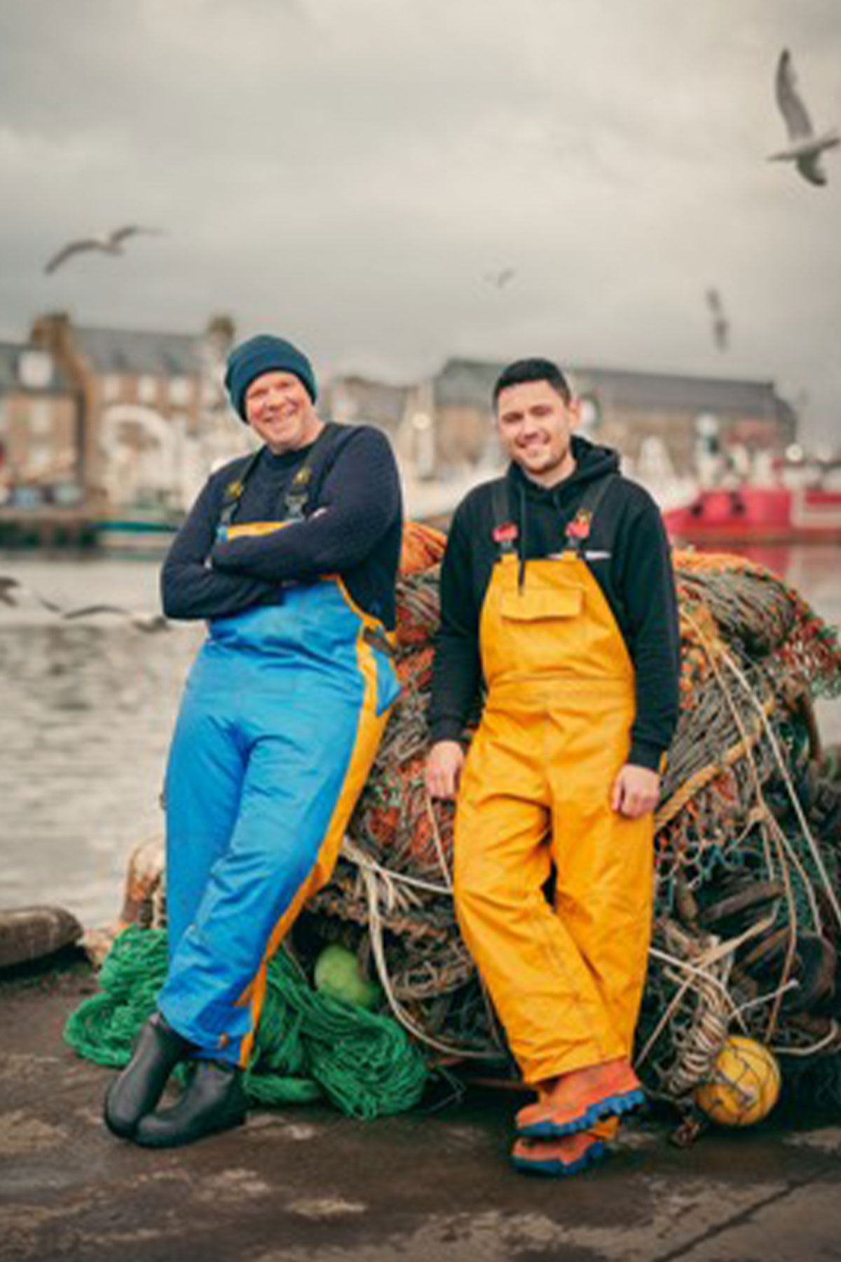Two people in colourful waterproof overalls stand smiling near a pile of fishing nets by a harbour. Overcast sky and seagulls create a coastal mood.