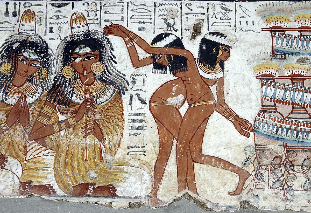 An ancient Egyptian mural showing a festive scene with musicians and dancers. Two seated women play flutes, dressed in gold robes and adorned with jewelry. To the right, two dancers move gracefully with raised arms. Hieroglyphics and decorated vessels appear in the background, adding to the lively atmosphere.