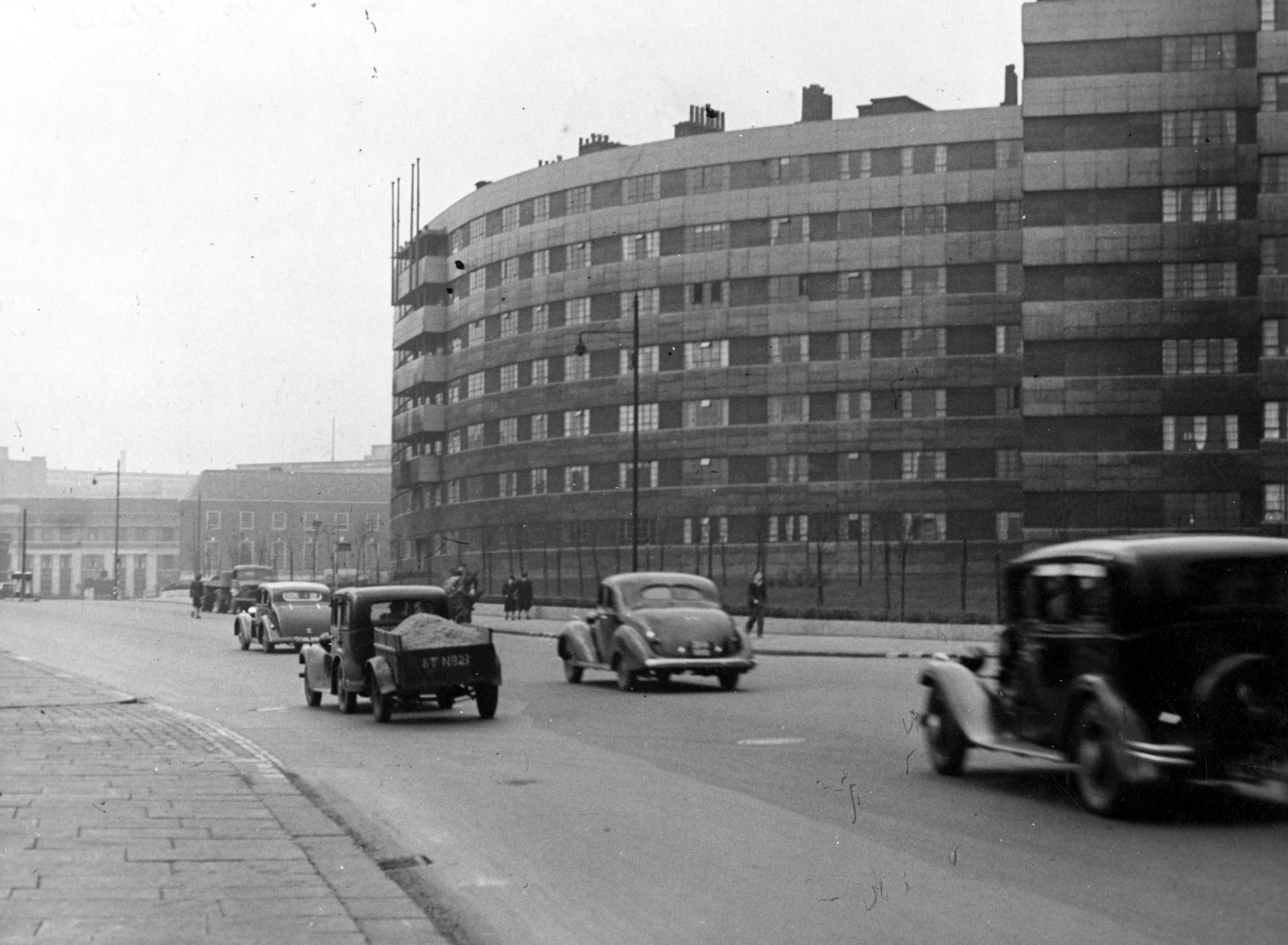 A large building of flats on the left hand side of the photo with a road running past them with cars on.