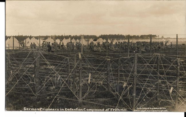 Photograph showing barbed wire fences around a British POW camp with tents and prisoners in the background.