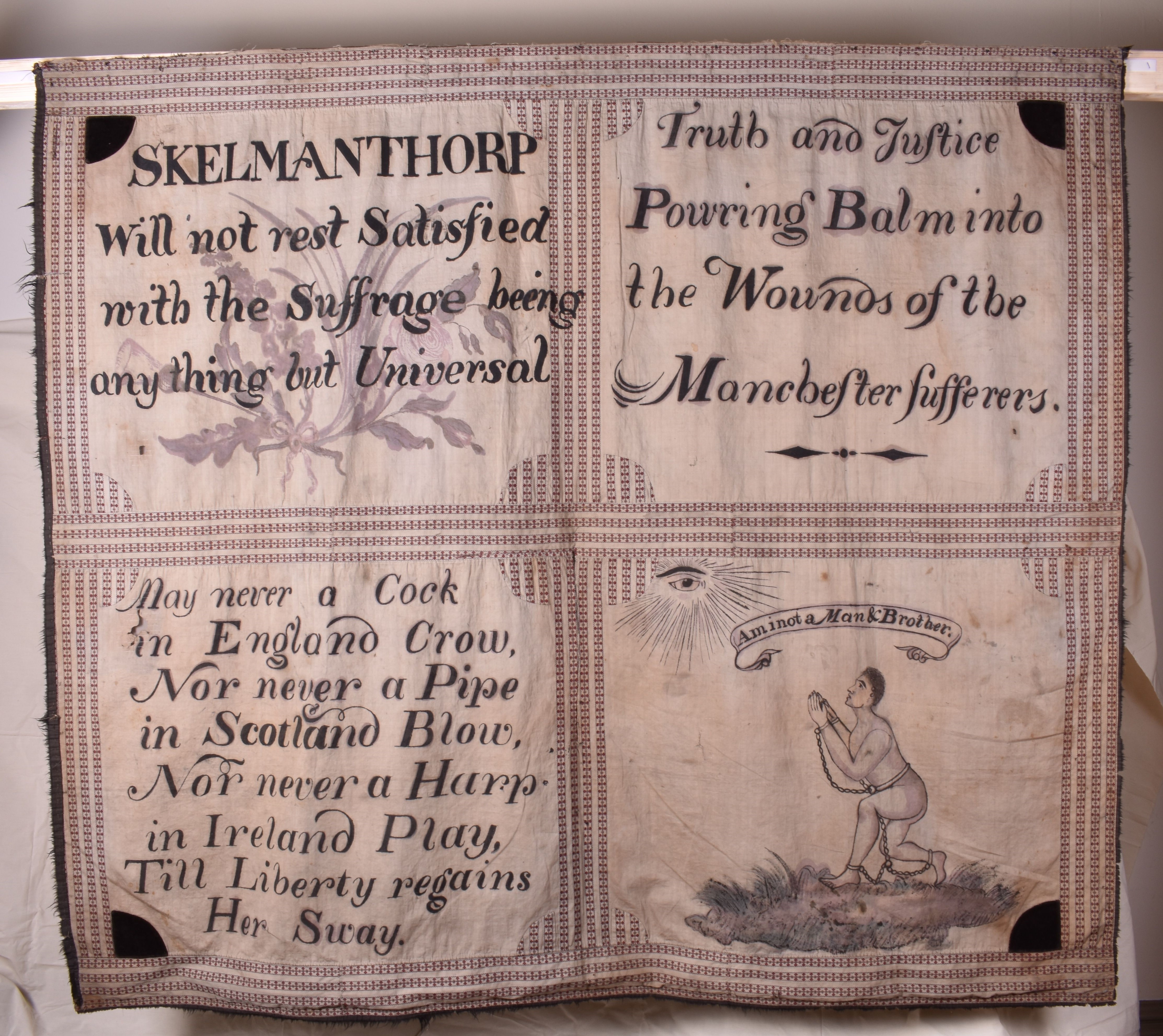 A historical protest banner from Skelmanthorpe, divided into four sections with text and illustrations. The top left section reads: 'SKELMANTHORP Will not rest Satisfied with the Suffrage being any thing but Universal'. The top right says: 'Truth and Justice Pouring Balm into the Wounds of the Manchester sufferers.' The bottom left section states: 'May never a Cock in England Crow, Nor never a Pipe in Scotland Blow, Nor never a Harp in Ireland Play, Till Liberty regains Her Sway.' The bottom right contains an illustration of an enslaved man in chains, kneeling with hands clasped in prayer beneath a radiant eye, with a banner above reading: 'Am I not a Man & a Brother.' The banner is hand-painted on cloth.