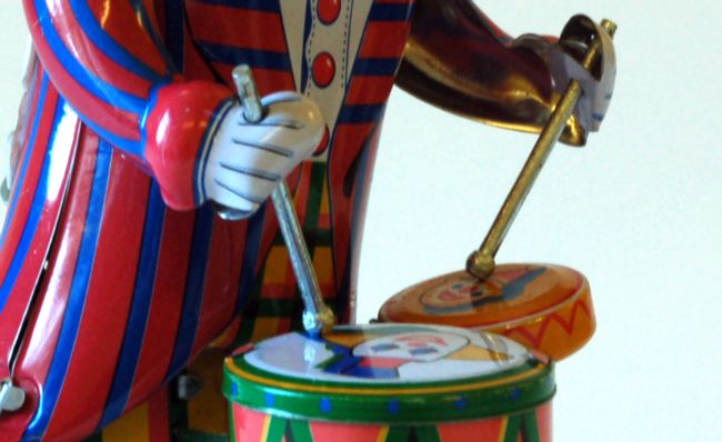 Close up view of toy clown  beating his drum.  he wears a blue and red stribed jacket and has two drums.