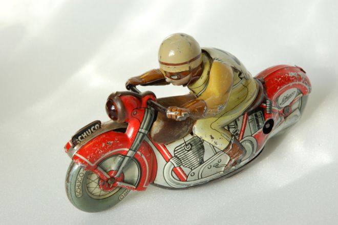 A 1950s tinplate, clockwork motorcycle.  The motorbike is red and the rider is wearing a yellow and brown outfit.  Some of the paint is missing.