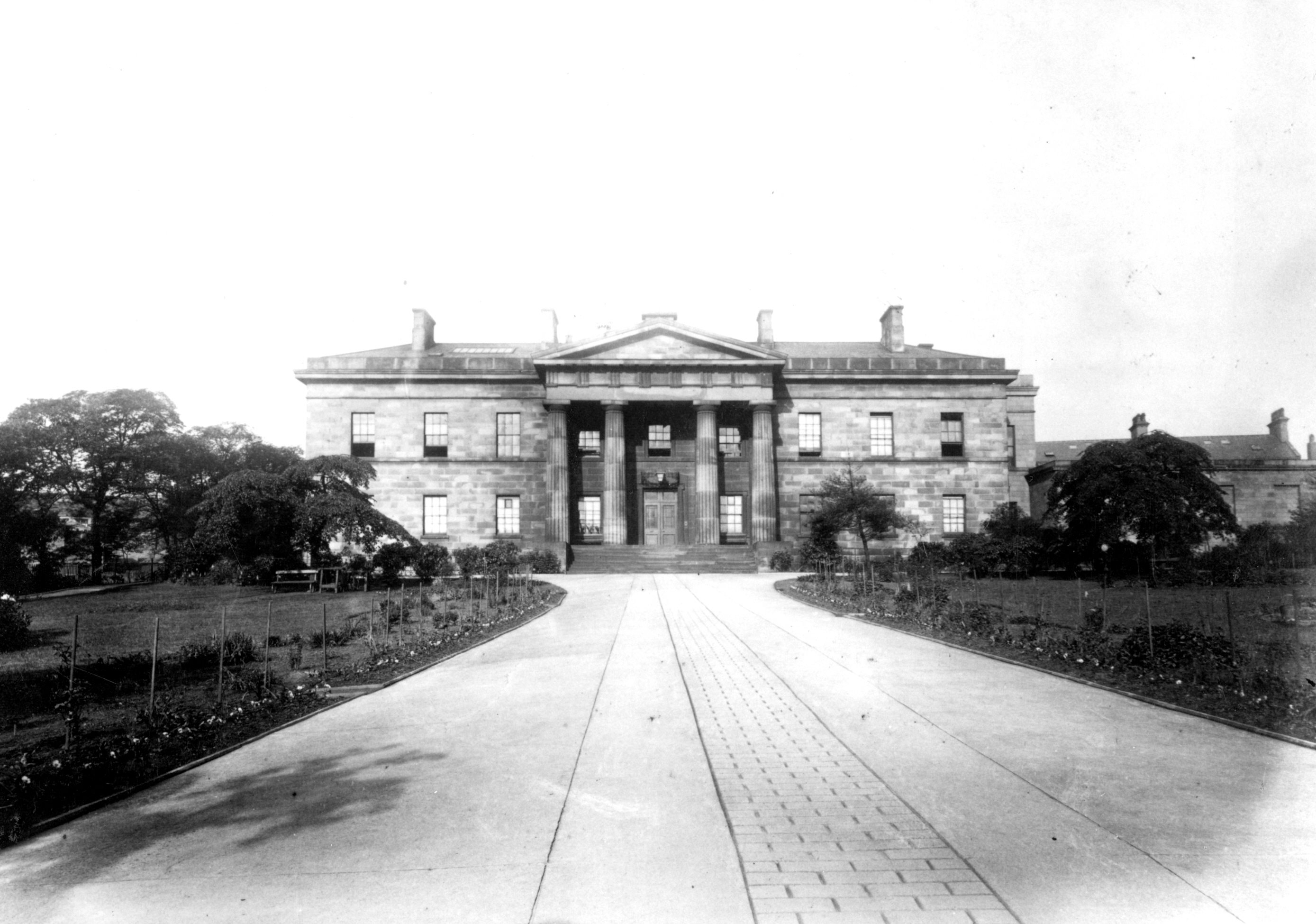 Black and white photograph of a grand neoclassical building with a columned portico and symmetrical facade. A wide, paved path leads to the entrance, flanked by manicured lawns and trees.