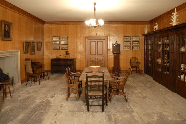 The Still Room, Temple Newsam House, Leeds showing a bare flagstone floor with a rough wooden table and chairs in the centre.  A longcase clock stands against one wall.