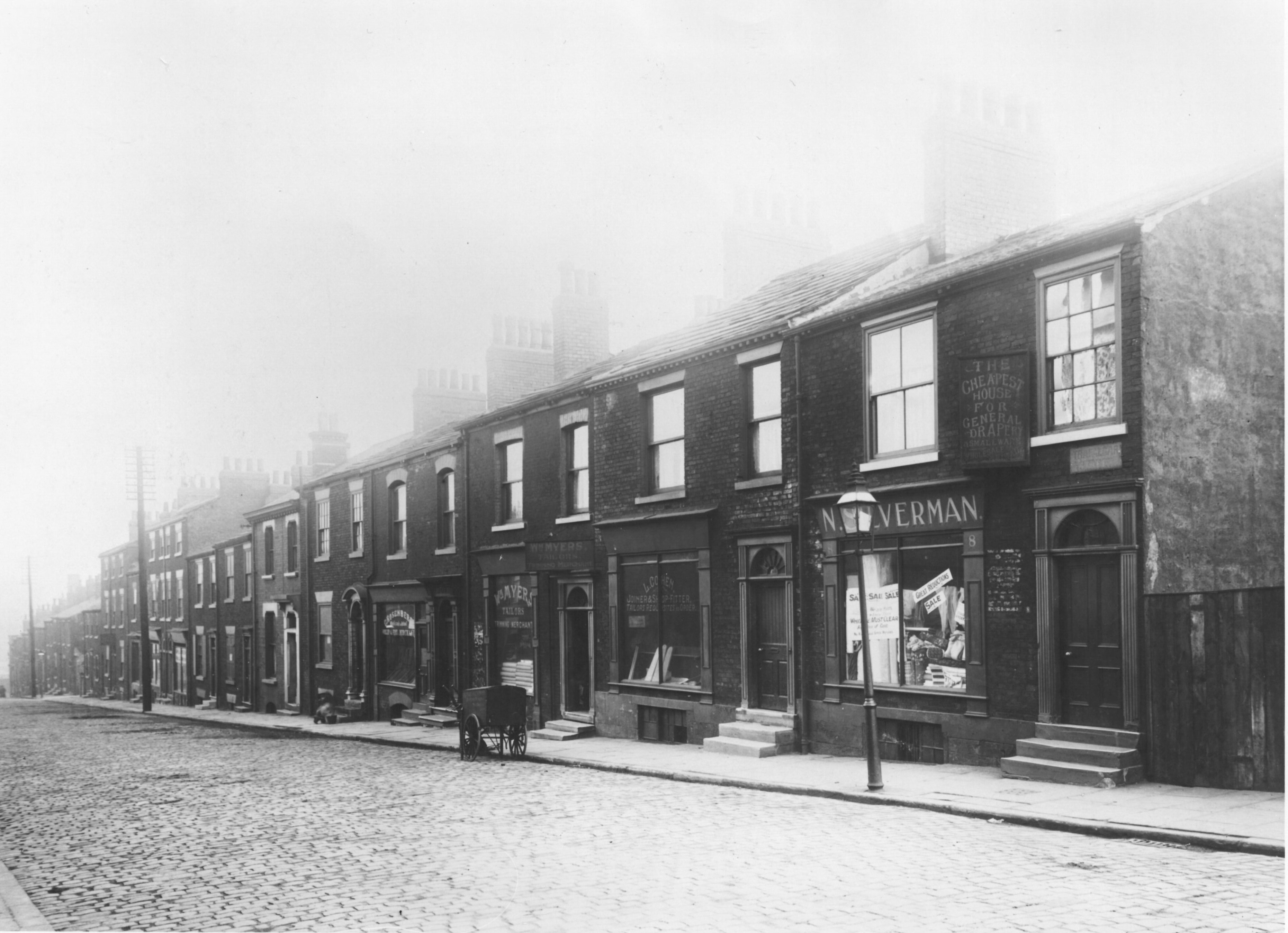 Black and white photo of a row of terraced houses and shops with cobbled street and gas lamps