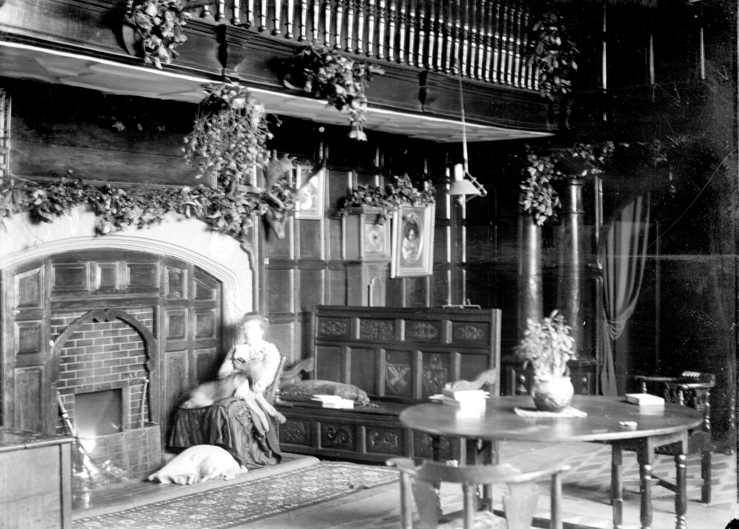 Black and white photo of a large, historic room with ornate wood panelling, fireplace adorned with greenery, a woman seated with pets, and a round table with a potted plant.