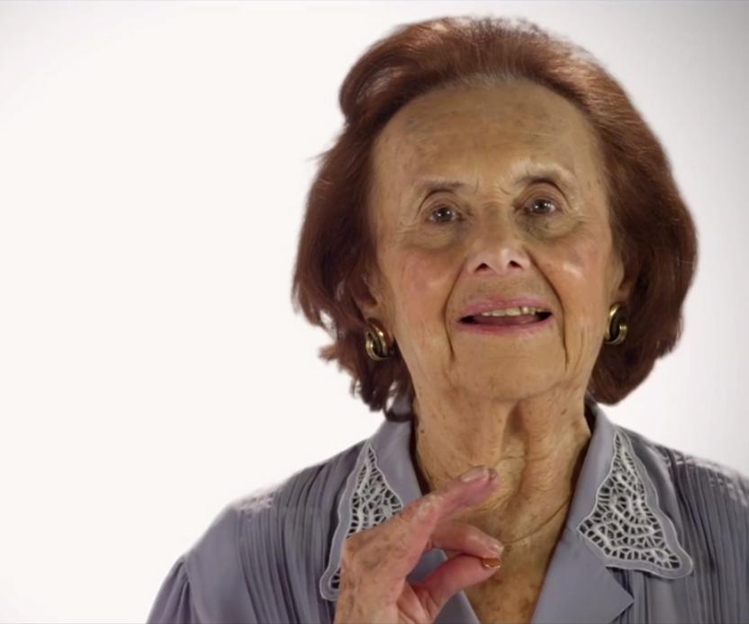 An elderly woman with short, light brown hair smiles softly, wearing a light gray blouse with lace details on the collar. She holds her hand up in a gentle gesture.