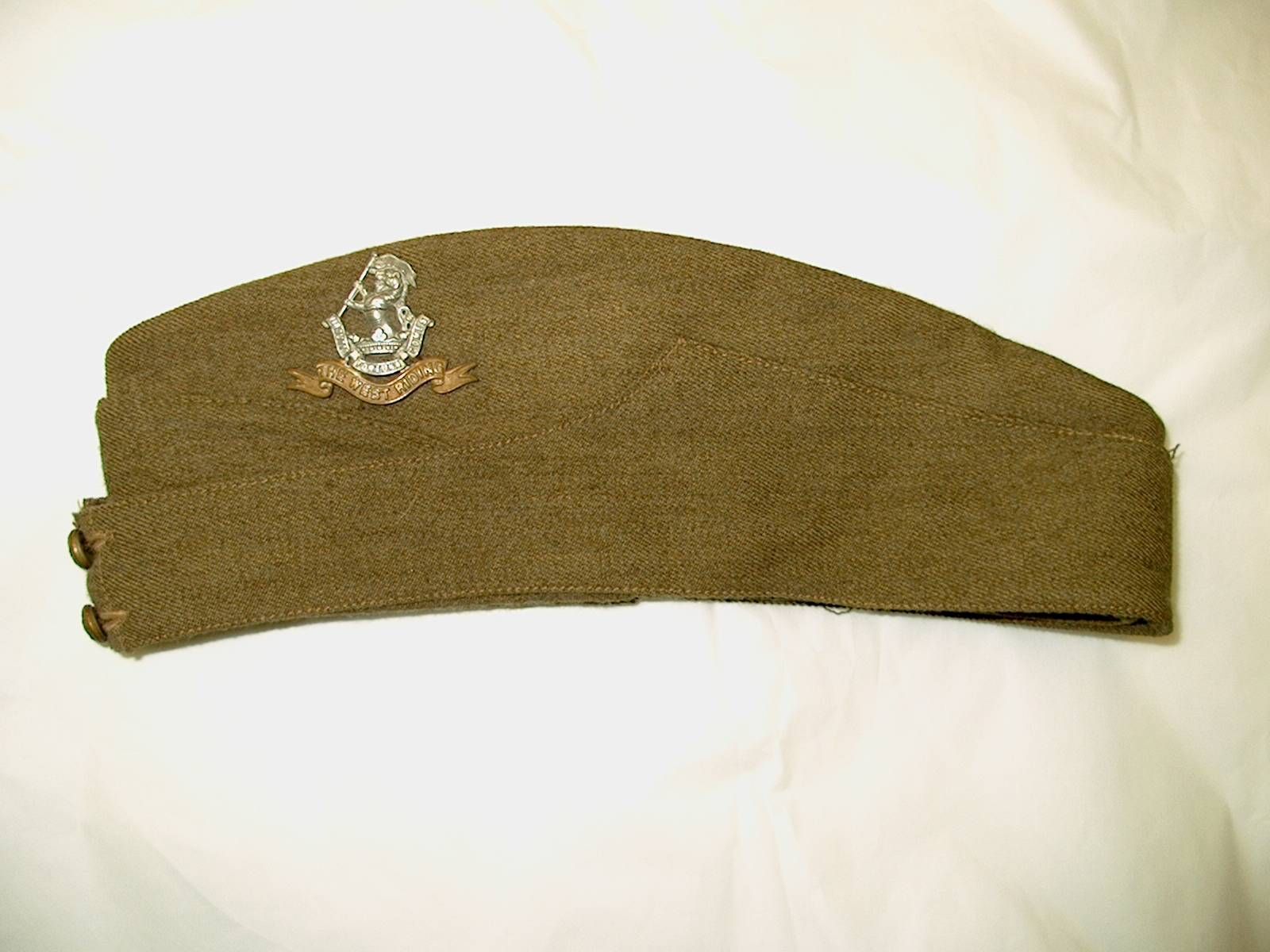 A brown woolen military side cap with a silver badge and emblem at the front, set against a plain white fabric background.