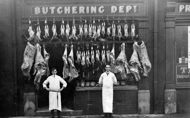 Two men in white aprons standing outside a butchers. There are many hides of meat hung up on hooks outside the shop.