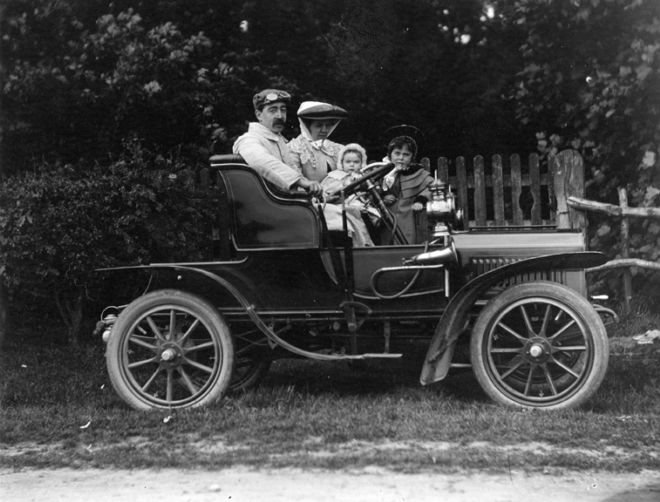 A family sitting in an early open top car.  The man and woman are wearing hats, and there are two children .  There is only one row of seat that takes up the width of the car.