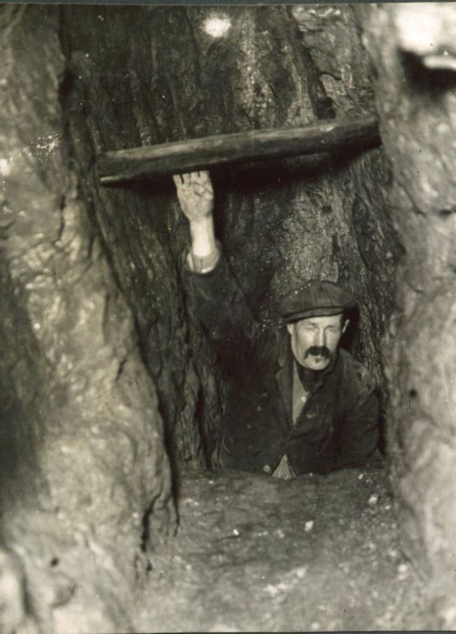 Black and white photograph of a man in a narrow shaft.  He is reaching up to grab a wooden strut