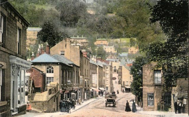 Coloured postcard of Matlock, showing a street lined with houses.  There is a horse and carriage on the street and people