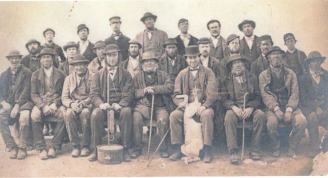 Black and white photograph of a group of men, the first two rows are seated, with a row of men standing behind.  Many are wearing bowler hats and three piece suits.
