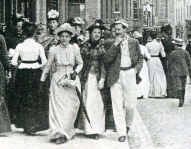 Black and white photo showing men and women walking down a road.  They are dressed in Victorian clothes